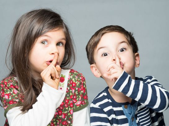 Zwei Kinder sitzen nebeneinander und halten den Finger vor den Mund um Stille anzudeuten.